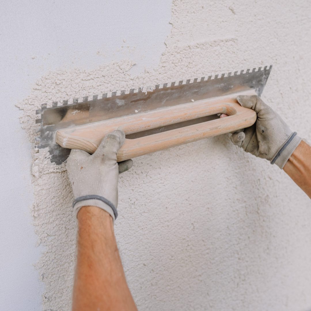 A worker smooths plaster on a wall using a notched trowel, focusing on an even finish. Close-up of construction work.